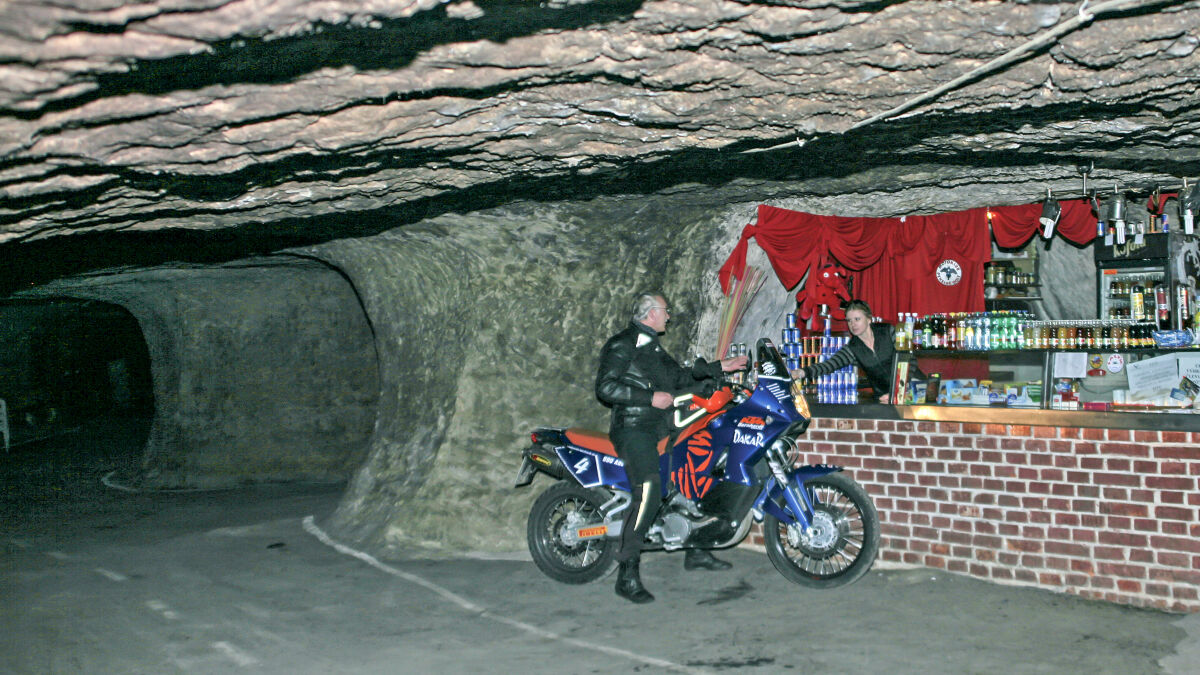 Stollen im Fels mit einer Bar aus Backstein, davor ein Mann neben einer blau-orangen KTM Stollen im Fels mit einer Bar aus Backstein, davor ein Mann neben einer blau-orangen KTM